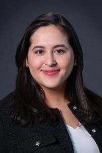 Kaitlynn smiles at the camera, wearing a professional black jacket in front of a dark background
