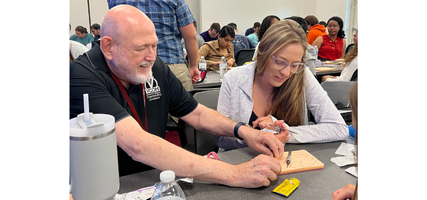 Professor shows student how to suture on wooden board with needle.