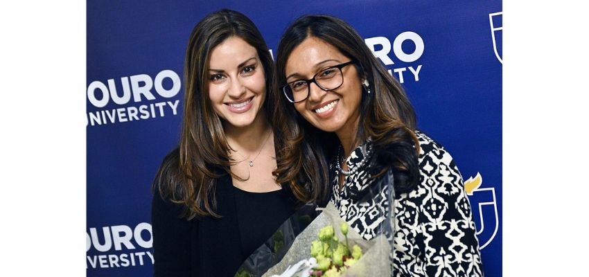 Two women smile together, one holding a bouquet of flowers, in front of a Touro University backdrop