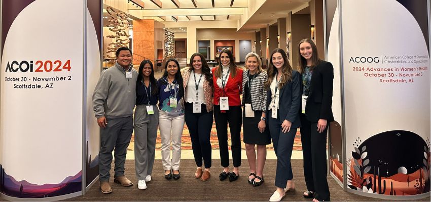 A group of students poses under a conference banner for ACOI 2024 and ACOOG 2024 in Scottsdale, Arizona.