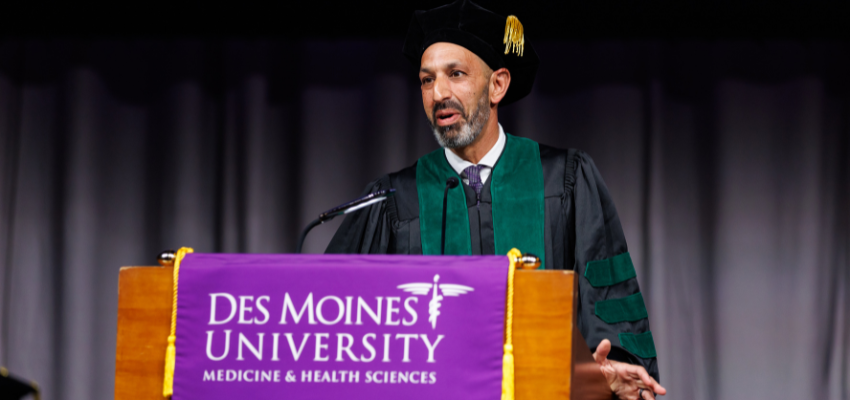 A speaker in academic regalia, including a black cap and green-trimmed gown, addresses the audience from a podium draped with a purple banner that reads 'Des Moines University Medicine & Health Sciences.'