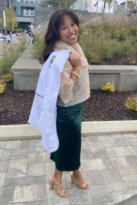 Medical student Camonayan standing outside with white coat over her shoulder. She is dressed elegantly in a green dress and beige heels, with a campus building in the background.