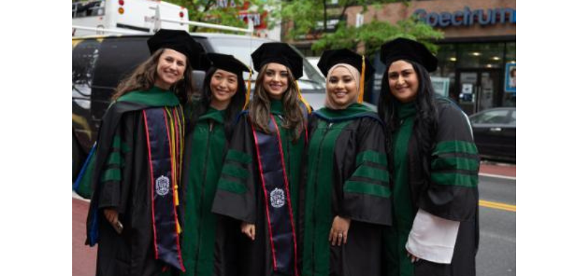 Five graduates from Touro College of Osteopathic Medicine pose together on a street in their black graduation gowns and caps, all smiling at the camera.