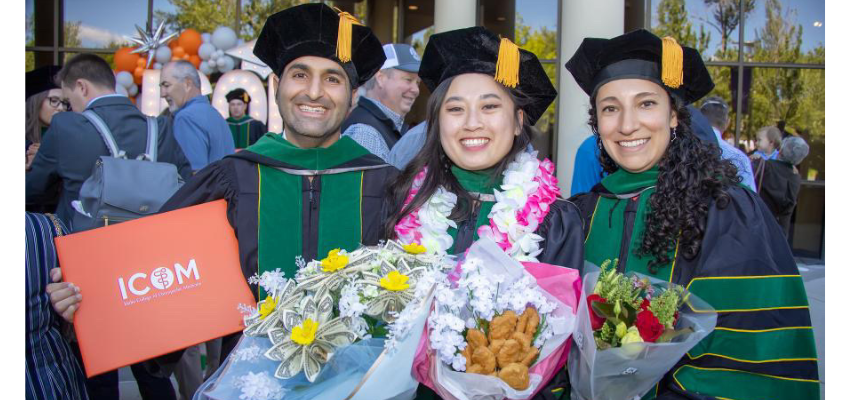 Three ICOM graduates in their caps and gowns holding bouquets and a diploma.