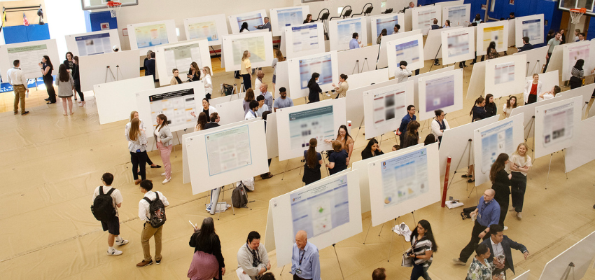 Wide view of a PCOM research poster exhibition with students and attendees.