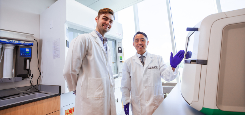 Two researchers in lab coats stand in a laboratory next to scientific equipment, smiling at the camera.