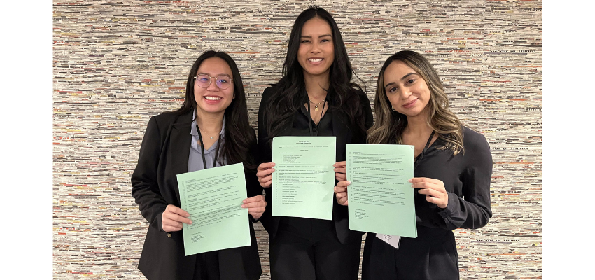 Three women in business attire hold up copies of the resolution, smiling, in front of a wall.