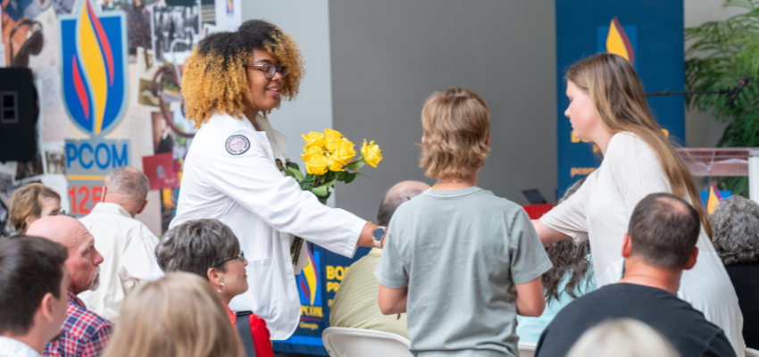 A medical student in a white coat hands a bouquet of yellow flowers to a young child at a community event.