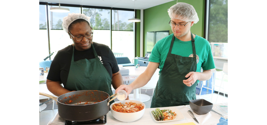 Two individuals wearing aprons and hairnets prepare a meal together in a kitchen setting