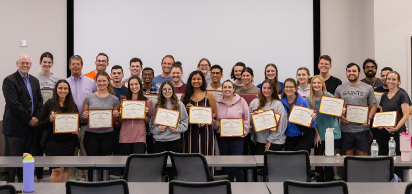 A large group of students hold certificates of achievement, standing together in a classroom with two instructors.