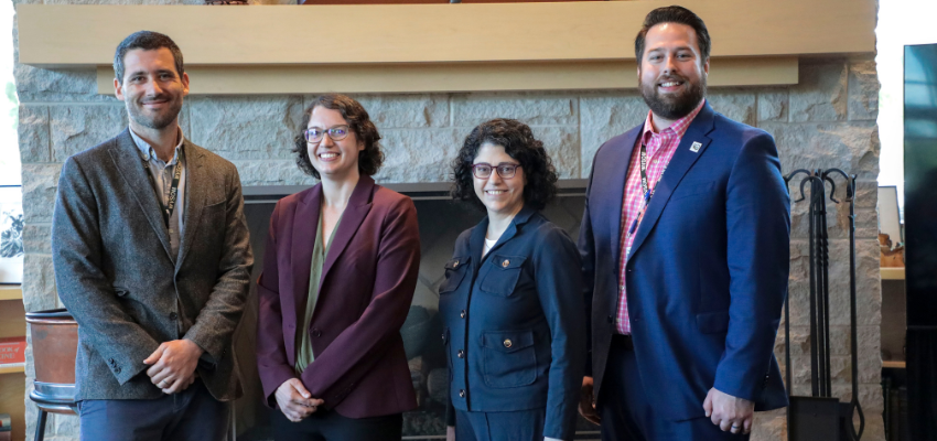 Four professionals in business attire stand together, smiling, in front of a fireplace in a formal setting.