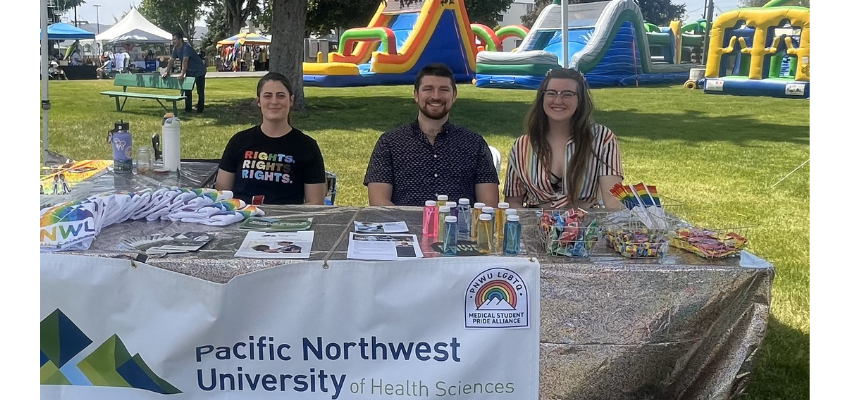 Three smiling students sit at a table filled with LGBTQ+ giveaways.