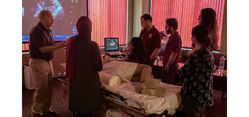 A group of medical students gathered around a patient simulator in a clinical skills lab. An instructor is explaining a procedure while students observe and interact with the simulation equipment.