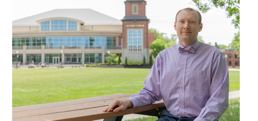 A man in a purple dress shirt and bow tie sitting on a bench outside with a university building in the background.