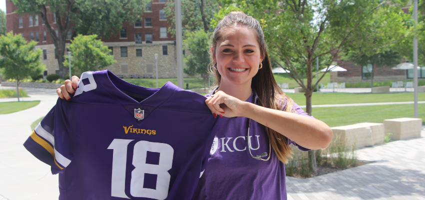 A student in a KCU shirt holding up a Vikings jersey outside on a sunny day.