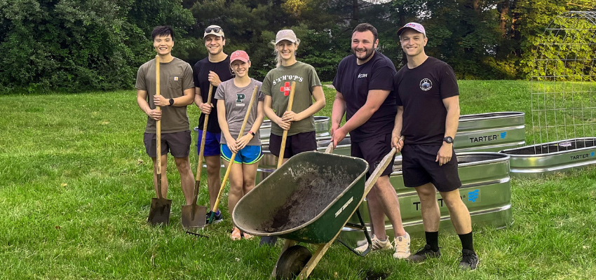 A group of six students outdoors, holding gardening tools and standing next to a wheelbarrow in a grassy area.