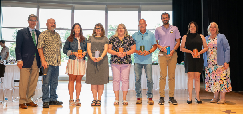 A group of eight individuals standing in a line on a stage, holding awards and smiling.