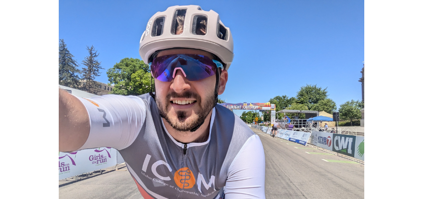 A man in a cycling helmet and sunglasses, wearing a jersey with the logo of Idaho College of Osteopathic Medicine, smiles at the camera while participating in a cycling event on a sunny day.