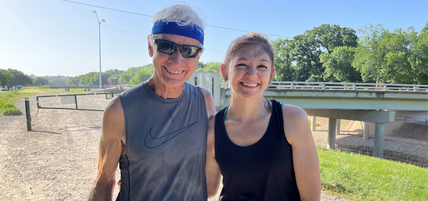 An older man with white hair, wearing sunglasses and a blue headband, smiles next to a younger woman with brown hair tied back, both dressed in athletic wear, standing outdoors on a sunny day near a bridge.