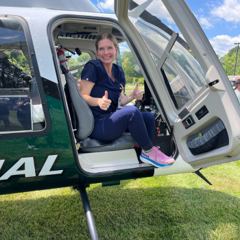 A smiling woman dressed in navy blue scrubs gives two thumbs up while sitting inside the open door of a helicopter with 