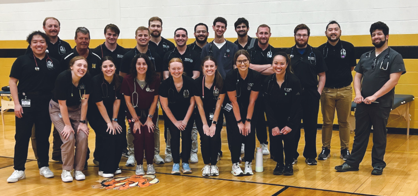 A group of medical students and instructors, wearing matching black polo shirts and stethoscopes, pose for a group photo in a gymnasium.