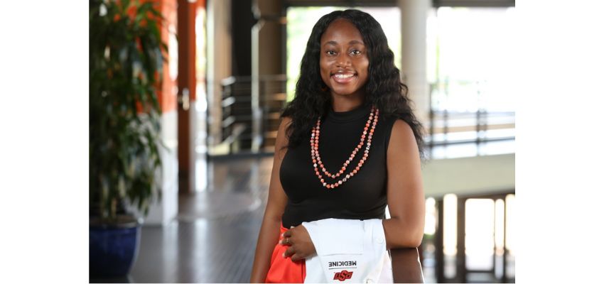 A smiling student holding her white coat in a university hallway.