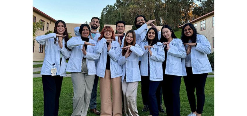 A group of TUCOM students in white coats form a 'T' with their hands while standing on a grassy campus.