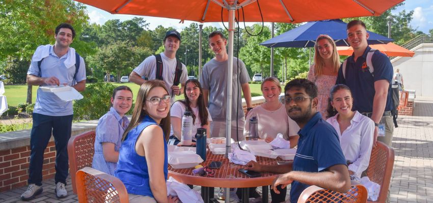 Group of students sitting around a table outside under umbrellas, enjoying a meal and smiling at the camera.