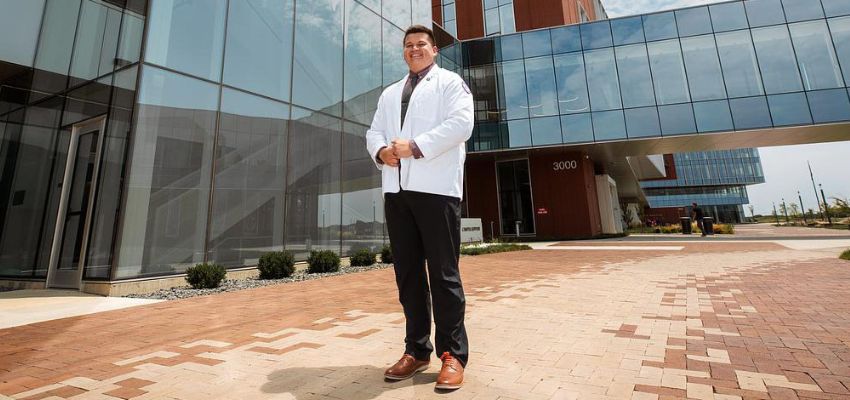 A medical student stands confidently in front of a modern healthcare building.