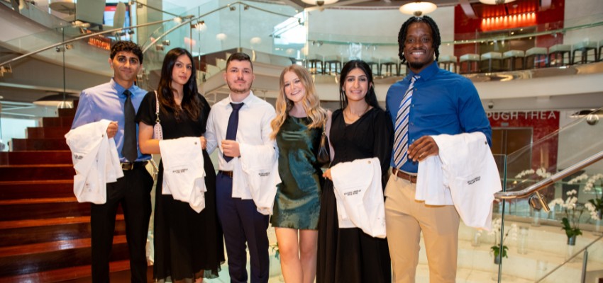 Six medical students holding white coats and smiling inside a building with a spiral staircase.