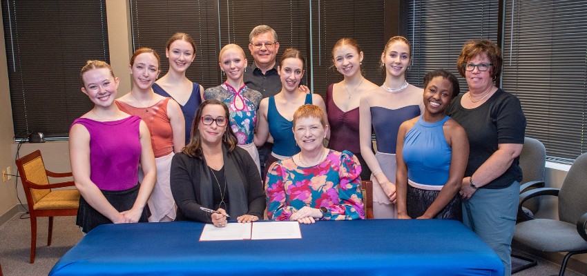 Group of ballet students and faculty smiling during a signing ceremony, with a blue table in front.