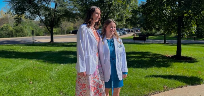 Two medical students posing in white coats outdoors, standing on a sunny day.