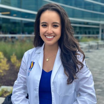 A smiling medical student wearing a white coat, standing outside a modern building.