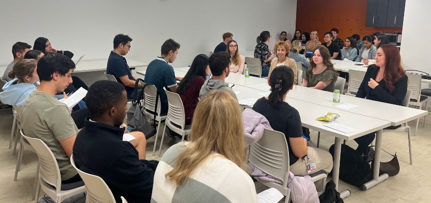 A classroom setting with students and faculty engaged in a discussion around a large table.