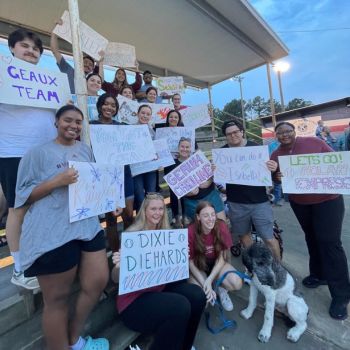 A group of students holding supportive signs, cheering at an outdoor event, with a dog present in the foreground.