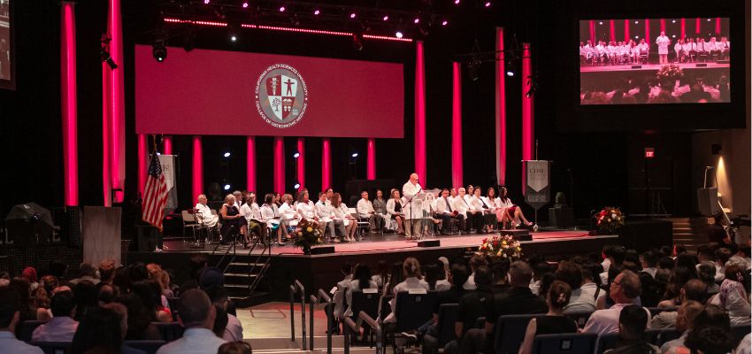 A formal ceremony at a medical school, featuring faculty and students on stage under a large school emblem.