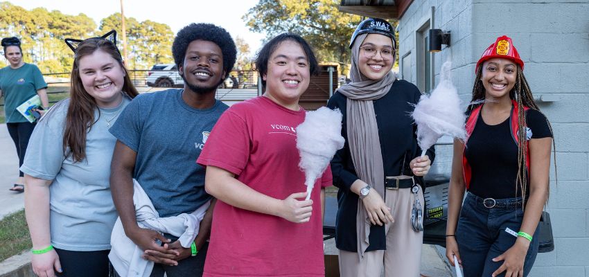 A group of five smiling young adults stands together outdoors, some holding cotton candy, with one wearing a firefighter hat and another with cat ears.