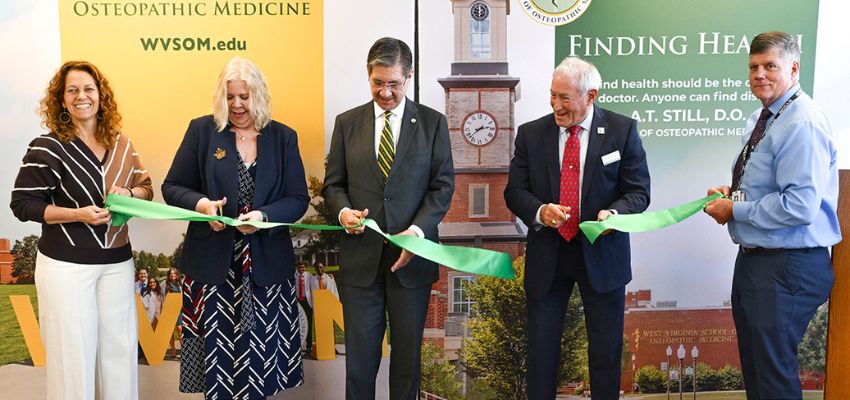 A ribbon-cutting ceremony with five people holding a green ribbon in front of banners for the West Virginia School of Osteopathic Medicine.