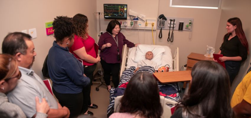 A group of medical professionals in a clinical setting, gathered around a simulated patient in a hospital bed, observing a presentation.