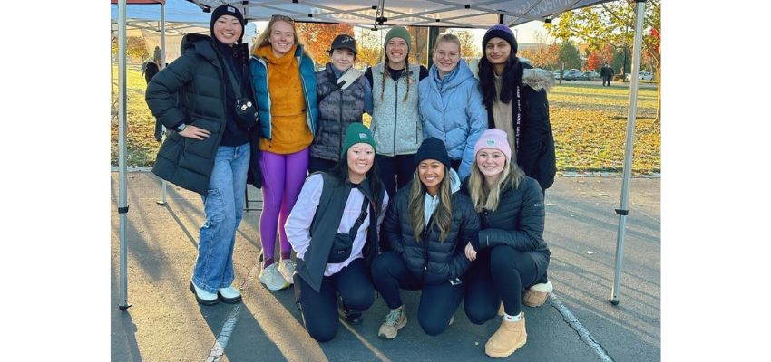 A group of students outdoors, dressed warmly, smiling under a tent on a clear autumn day.