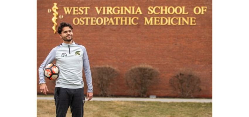 A man holding a soccer ball standing outside the West Virginia School of Osteopathic Medicine building.