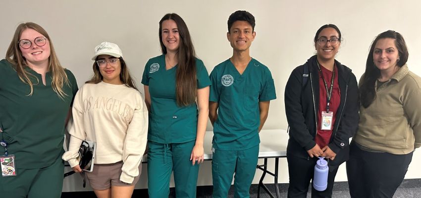 A group of students in scrubs and casual clothing smiling and standing together in a classroom.