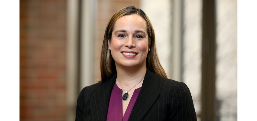 A professional woman in a blazer and blouse smiling in a well-lit office setting.