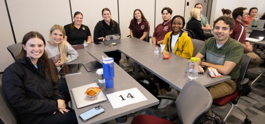 Students sitting around a table in a classroom, smiling and engaged in a discussion.