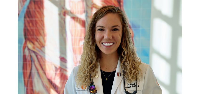 A smiling medical student in a white coat poses in front of a brightly lit, colorful background.