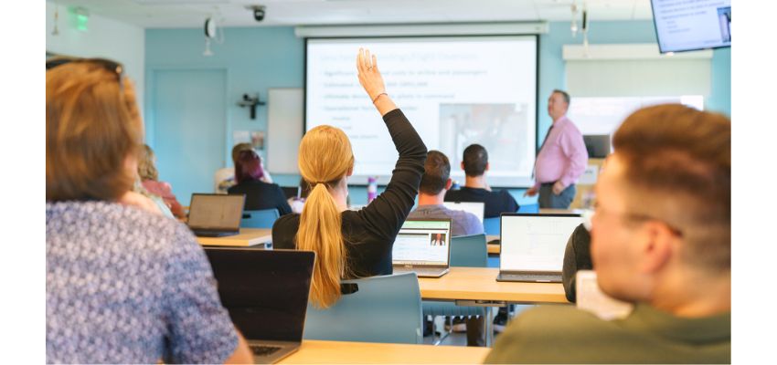 A classroom setting where a student raises their hand while others work on laptops during a lecture.