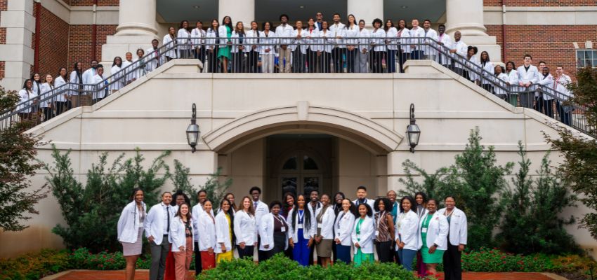 A large group of medical students in white coats gather outside a university building for a group photo at VCOM-Louisiana.