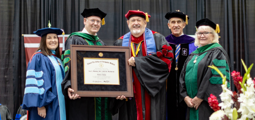 Five people in academic regalia are on stage during a graduation ceremony. One person in the center is holding a framed diploma and smiling, while the others stand proudly beside them.
